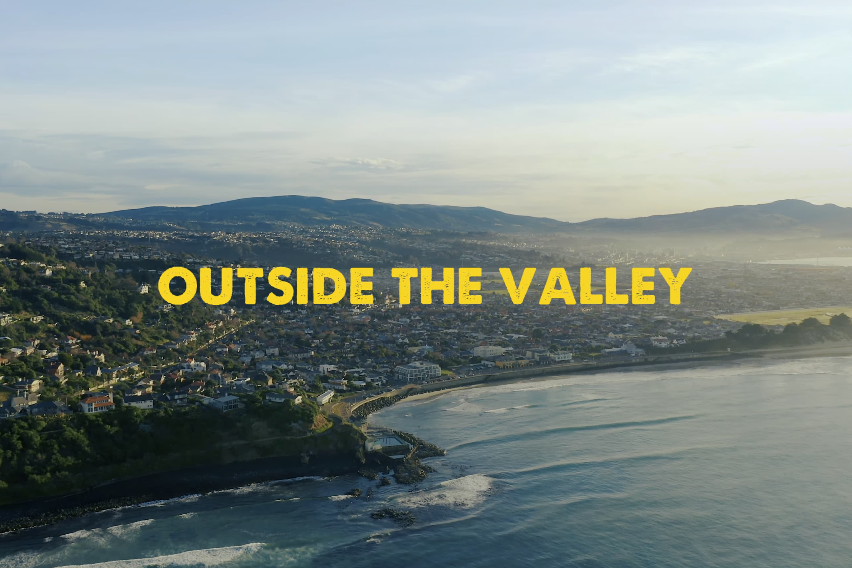 Outside the valley movie logo over a drone photo of Dunedin